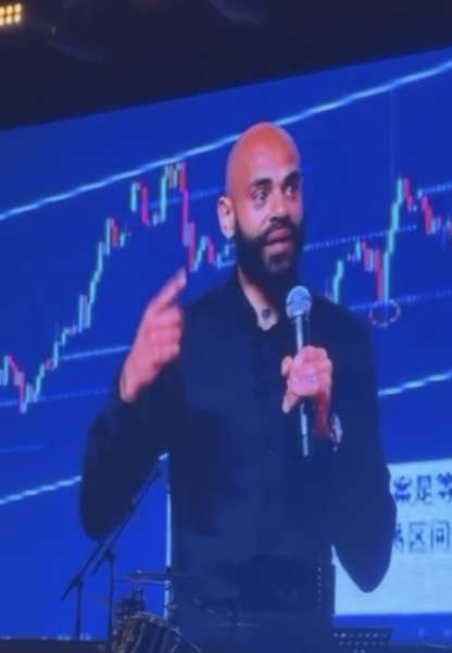 Ken Chigbo speaking on stage with trading charts